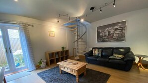 Una sala de estar con una mesa de centro de madera, un sofá negro, una escalera de caracol y un cuadro con una vista urbana en la pared.