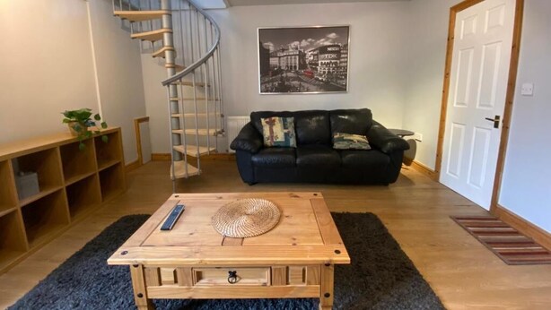 Una sala de estar con una mesa de centro de madera, un sofá de cuero negro, una escalera de caracol y un cartel con un paisaje urbano en la pared.