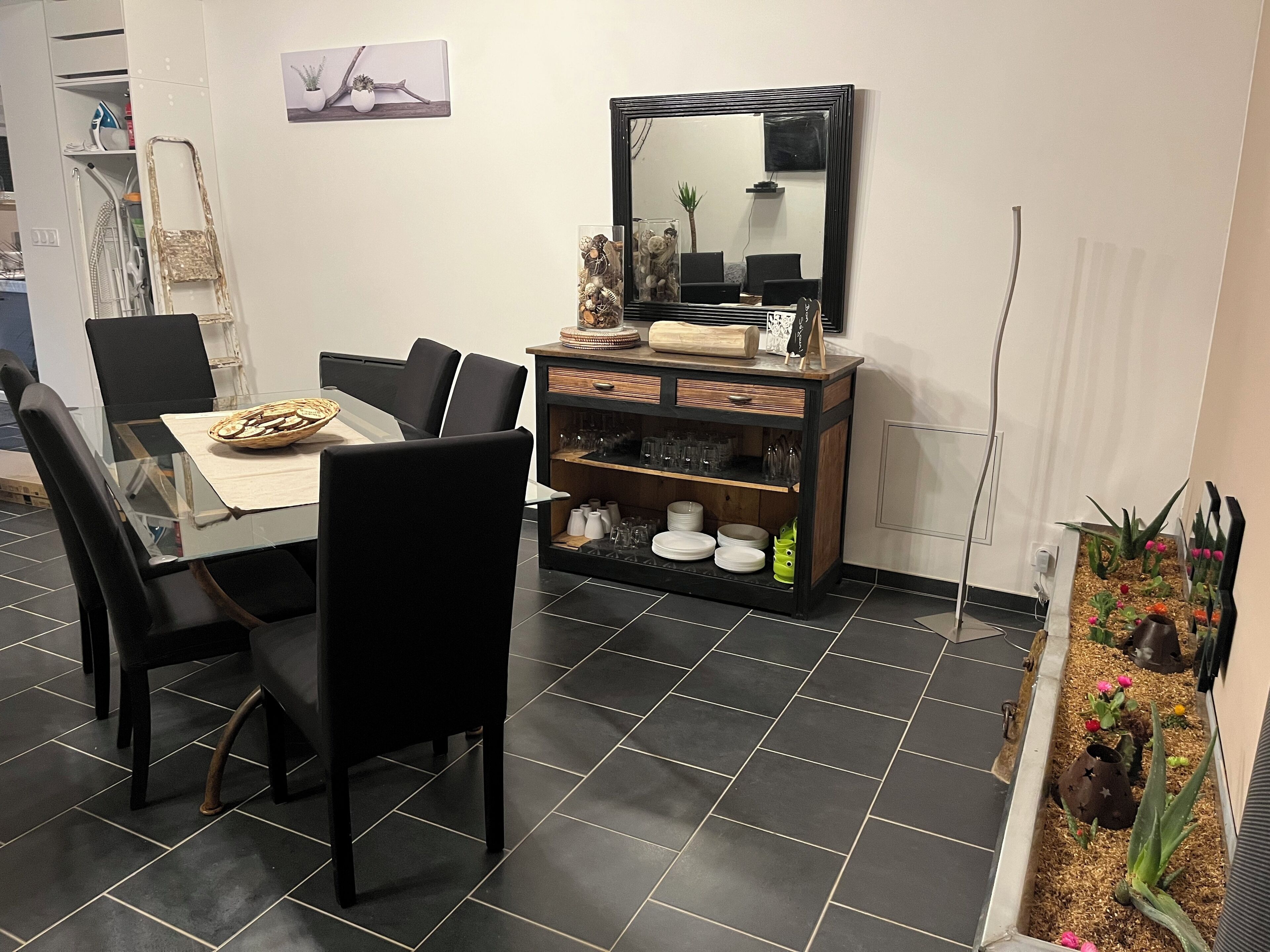 A dining area with a glass table, black chairs, a wooden sideboard with glassware, and a mirror.