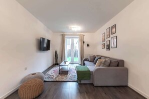 A modern living room with a grey sofa, a flat-screen TV mounted on the wall, a patterned rug, and a small round ottoman. - Host & Stay | Moresby Way (Peterborough)