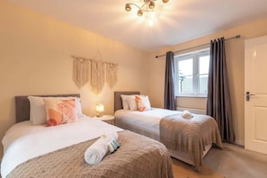 A hotel room with two beds, a window with curtains, a wall-mounted macrame decoration, and a ceiling light fixture. - Host & Stay | Moresby Way (Peterborough)