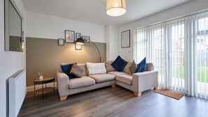 A modern living room with a grey sectional sofa, a small side table, a floor lamp, and a large window with a view of a backyard. - Host & Stay | Hewitt Close (Peterborough)