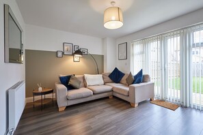 Ein modernes Wohnzimmer mit einem grauen Ecksofa, einem kleinen Beistelltisch, einer Stehlampe und einem großen Fenster mit Blick auf einen Garten.