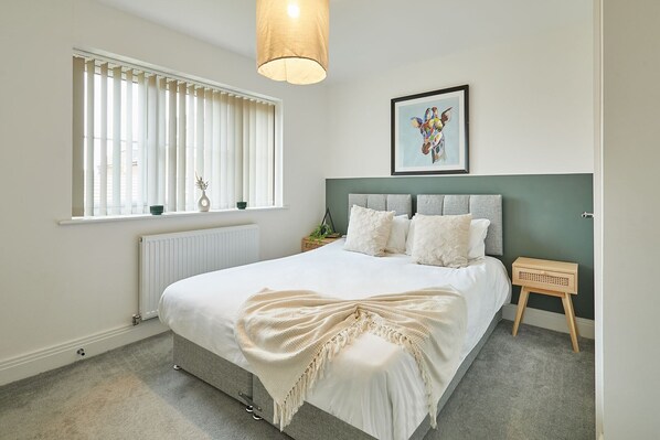 Ein Schlafzimmer mit Bett, Fenster mit Jalousien, Heizkörper, Nachttisch und einem gerahmten Bild an der Wand.