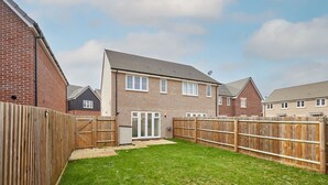 A residential backyard with a wooden fence, a grassy lawn, and a house with a brick exterior and a tiled roof. - Host & Stay | Hewitt Close (Peterborough)