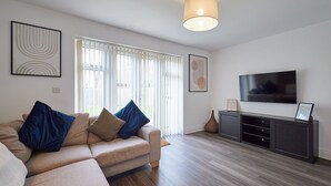 A modern living room with a beige sofa, blue cushions, a flat-screen TV, and a wooden floor. - Host & Stay | Hewitt Close (Peterborough)