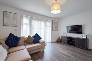 Un salon moderne avec un canapé beige, des coussins bleus, un téléviseur à écran plat et un plancher en bois.