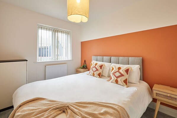 Ein Schlafzimmer mit Bett, einem Fenster mit Jalousien, einem Heizkörper, einem Nachttisch mit Lampe und einer orangefarbenen Akzentwand.