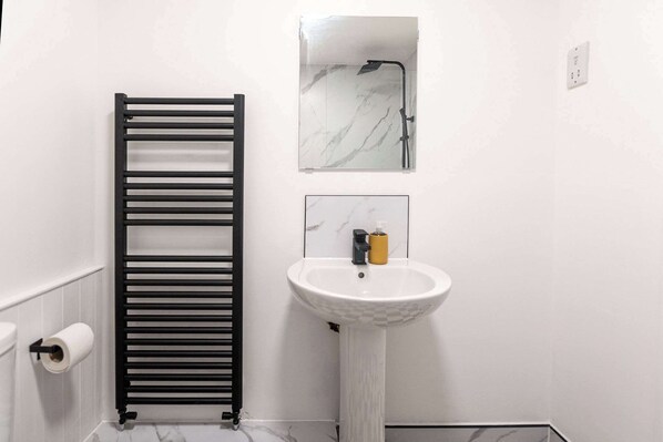 Ein Badezimmer mit einem schwarzen Handtuchhalter, einem weißen Waschbecken mit Seifenspender und einem Spiegel mit Duschkopf darüber.