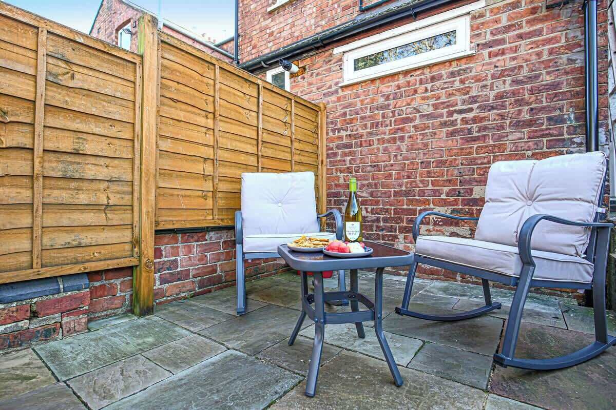 An outdoor patio with a wooden fence, two white cushioned chairs, a table with a bottle and plate of food, and a brick wall.