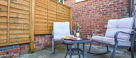An outdoor patio with a wooden fence, two white cushioned chairs, a table with a bottle and plate of food, and a brick wall.
