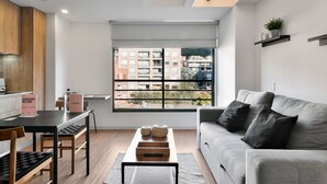Een moderne woonkamer met een grijze bank, zwarte kussens, een houten salontafel en een uitzicht op een stadsgezicht door grote ramen.