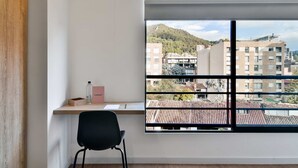 Een kamer met een groot raam, een bureau met stoel, en uitzicht op gebouwen en een heuvel.