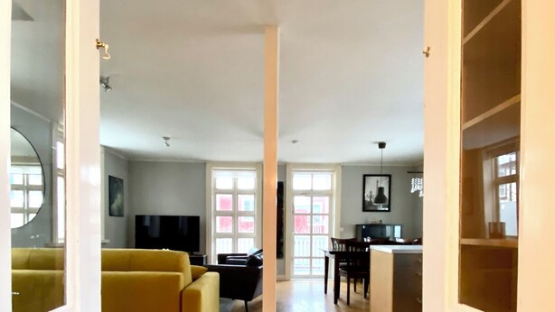 A spacious living area with a yellow sofa, a black chair, and a dining table with chairs. There is a large window with pink curtains, and a mirror on the left wall. - Day Dream Vesturgata 5a (Reykjavik)