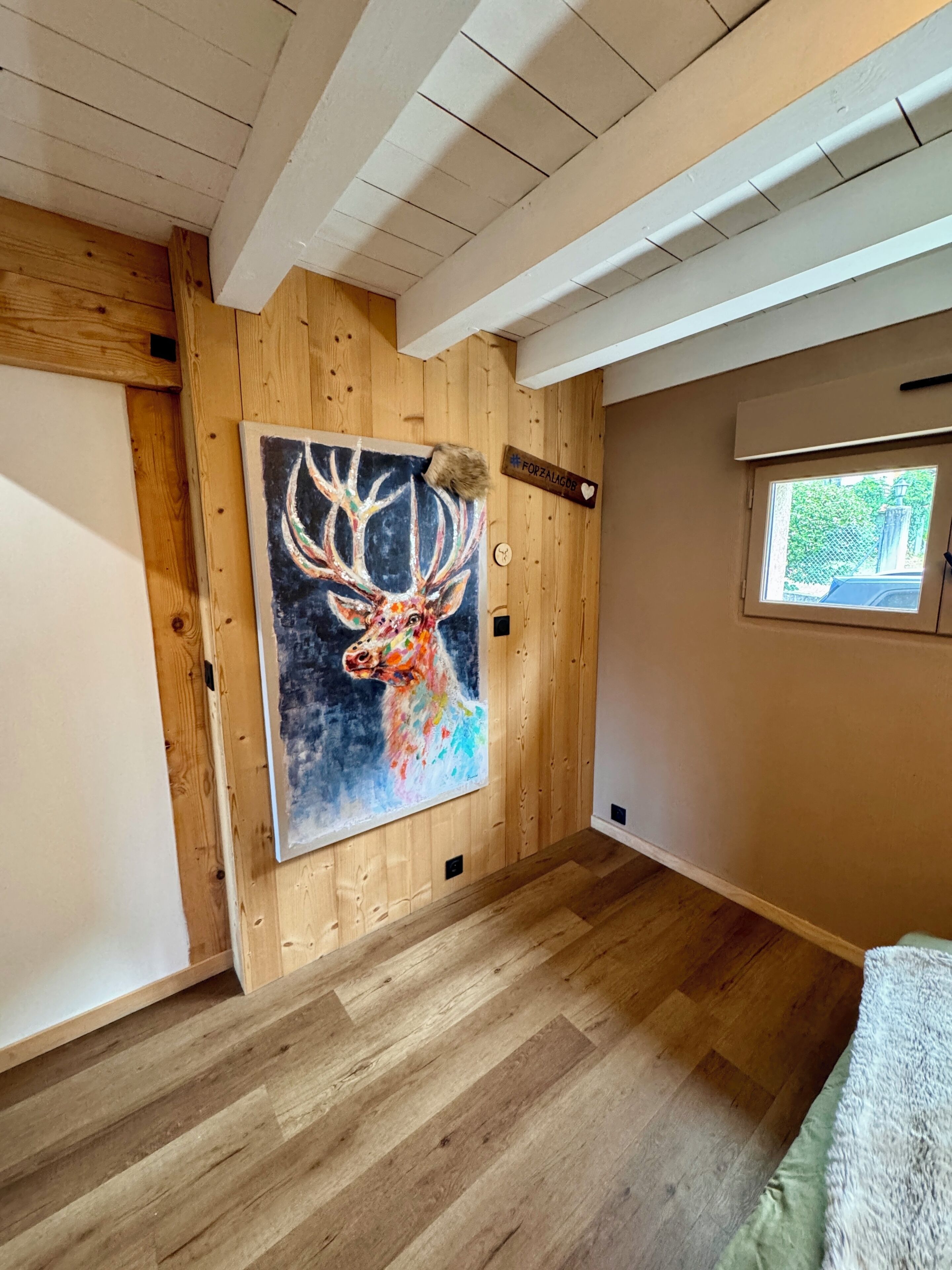 Ein Zimmer mit Holzwänden, einem großen Bild eines Rehs, einem Fenster mit Blick und einem Bett.
