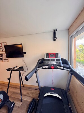 A treadmill in a room with a television on the wall, a small stool, and a window with a view.