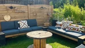 Een terras met een houten wand, een ronde houten tafel en een bank met kussens.