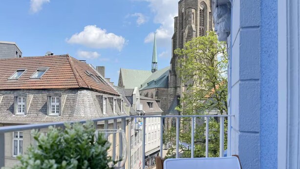 A balcony with a potted plant, a chair, and a view of a church with a spire.