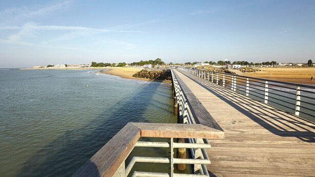 Un muelle de madera se extiende sobre aguas tranquilas, con una playa arenosa y edificios a lo lejos.