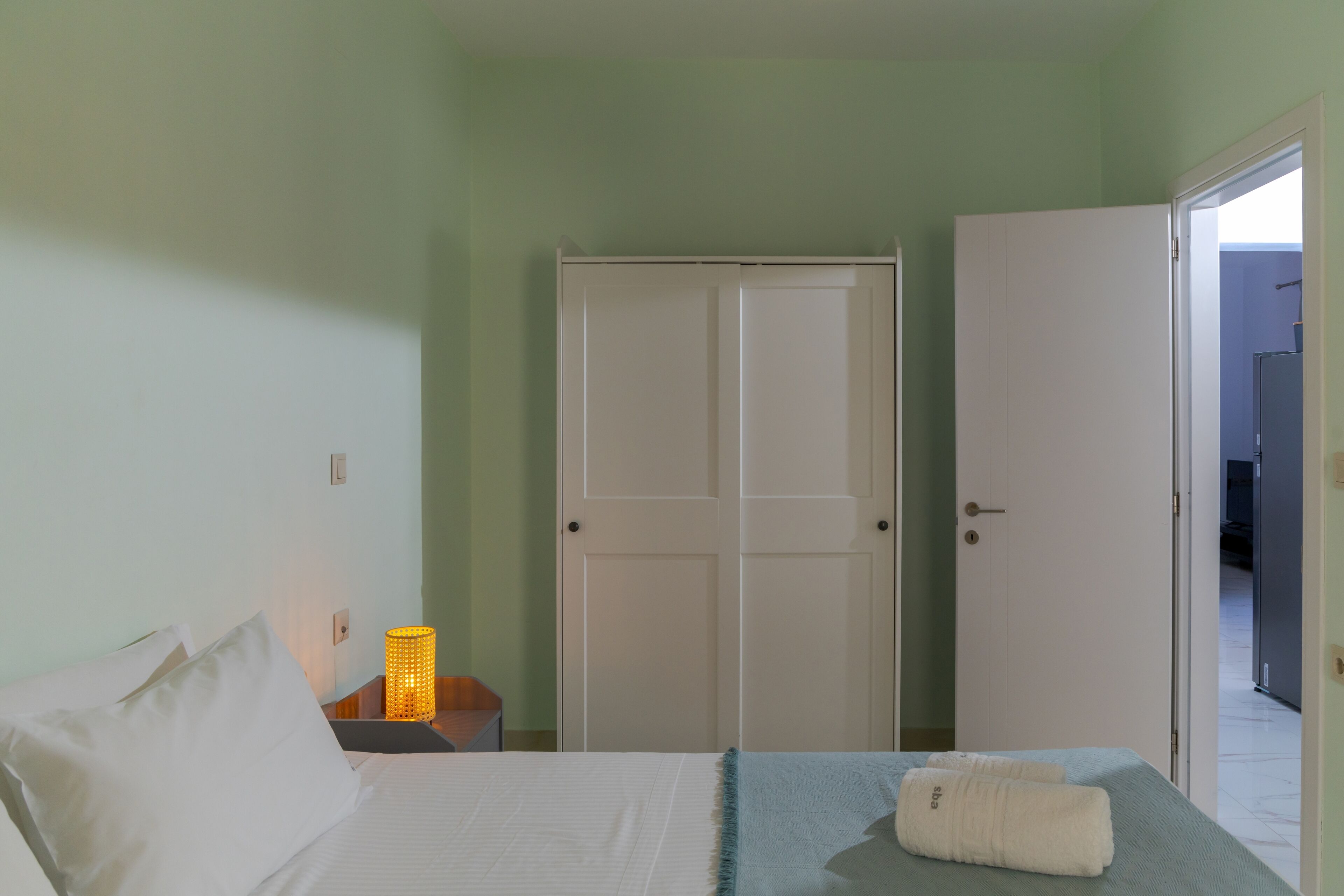 A bedroom with a bed, a nightstand with a lamp, a wardrobe, and an open door leading to another room.