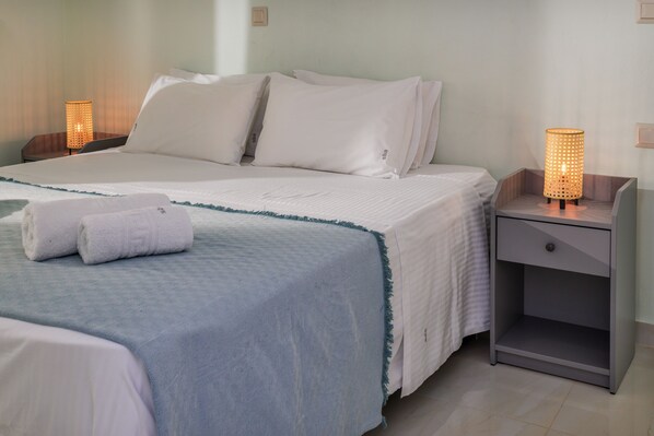Una cama bien hecha con ropa blanca y un detalle azul, una mesita de noche con una lámpara y un tocador con un cajón.