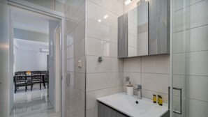 A modern bathroom with a white sink, mirror, and tiled walls. There is a door leading to a dining area with a table and chairs.