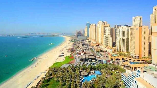 A coastal cityscape with high-rise buildings, a beach, and a resort area with pools and greenery.