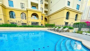 A swimming pool with clear blue water, surrounded by a yellow building with balconies and a fence.