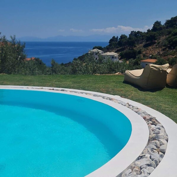 A swimming pool with a curved edge, surrounded by a stone border, and a view of the sea and hills in the background.
