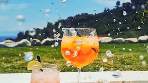 Dos vasos de bebida naranja sobre una superficie blanca, con una piscina y vegetación al fondo.