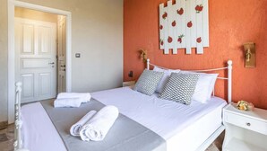 Una cama bien tendida con sábanas blancas y toallas enrolladas, una mesita de noche blanca con un pequeño jarrón, y una pared decorada con motivos frutales.