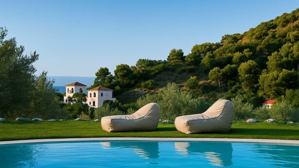 A pool with two beige lounge chairs, a grassy area, and a building with a red roof in the background. - Skopelos Seaview Suite - Demeter Summer Nest (Stafylos)