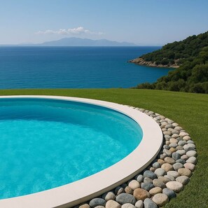 A swimming pool with a clear blue water, surrounded by a stone border and a grassy area, with a view of the sea and distant mountains.