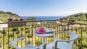 Ein Balkon mit Tisch und zwei Stühlen, der auf eine Küstenlandschaft mit Grünflächen und Gebäuden blickt.
