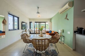 A dining area with a round table and wicker chairs, a kitchen with a coffee machine, and a view of greenery through large windows.