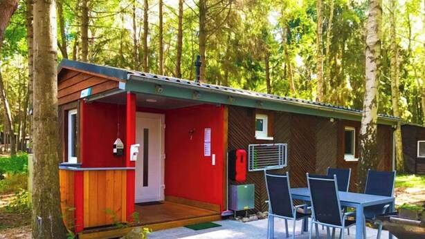 Une petite cabane à l’extérieur rouge, avec une terrasse en bois, une table et des chaises, et un chemin en pierre.