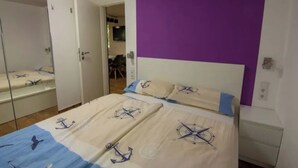A bedroom with a bed featuring nautical-themed bedding, a purple accent wall, and a white headboard. - Meiks Ostsee-Hütten - Holiday home 5 (Dranske)