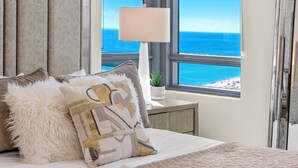 Una cama bien hecha con un cubrecolchón acolchado, un almohadón blanco mullido y almohadas decorativas con un estampado geométrico. Se ve una mesita de noche con una lámpara y una planta pequeña. La habitación tiene un ventanal grande que ofrece vistas al océano.