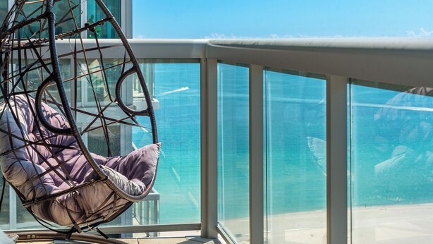 Una silla de mimbre con cojines junto a una barandilla de cristal con vistas a la playa.