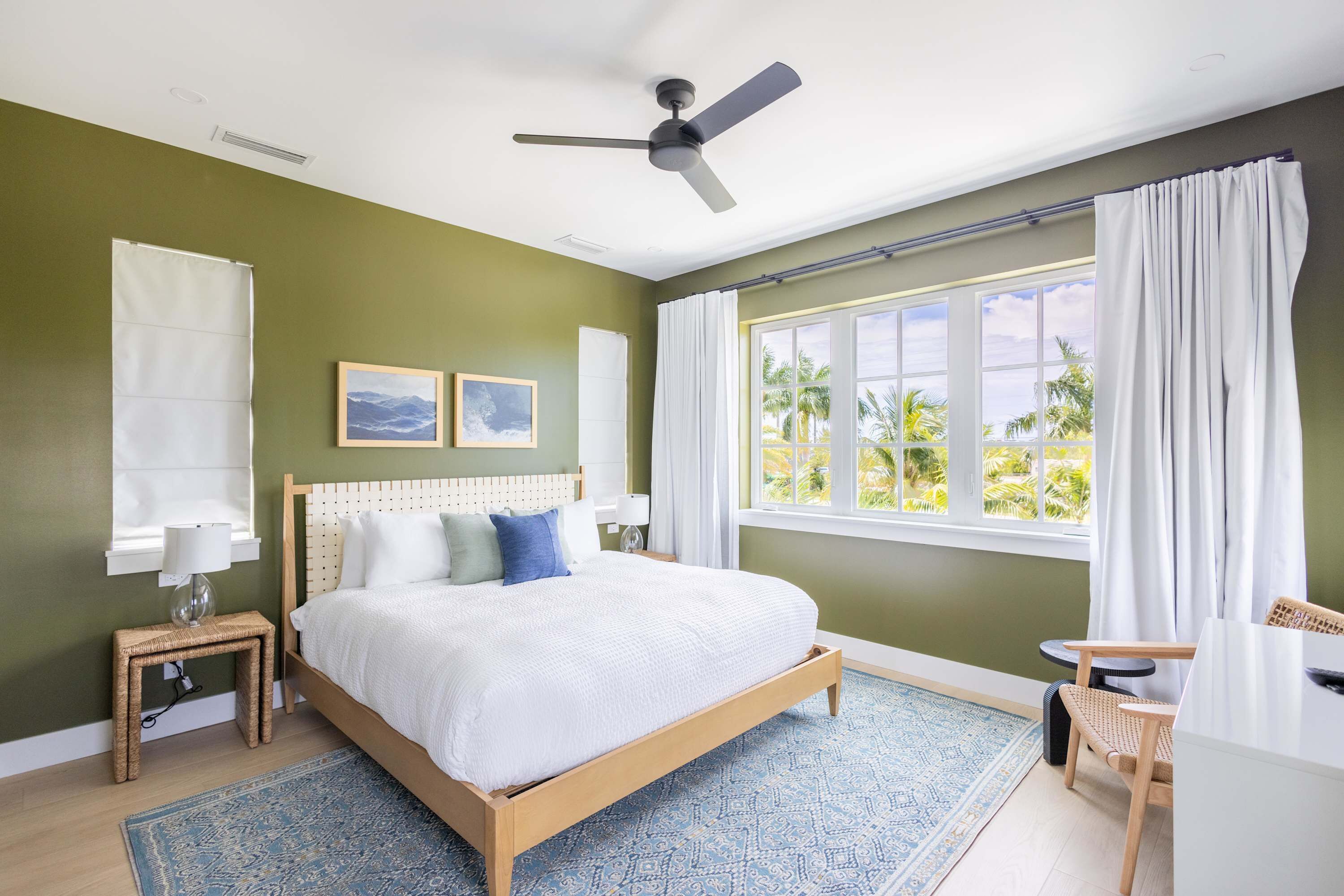A bedroom with a bed, a ceiling fan, a window with curtains, a bedside table with a lamp, and a small chair.