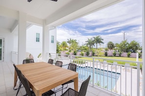 A covered patio with a wooden table and chairs, a pool, and a view of a residential area with palm trees. - Palmgale Estate at Lunara Bay (Key West)