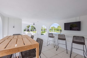 A spacious outdoor area with a wooden table and wicker chairs, a TV mounted on the wall, and a view of a garden through arched openings.