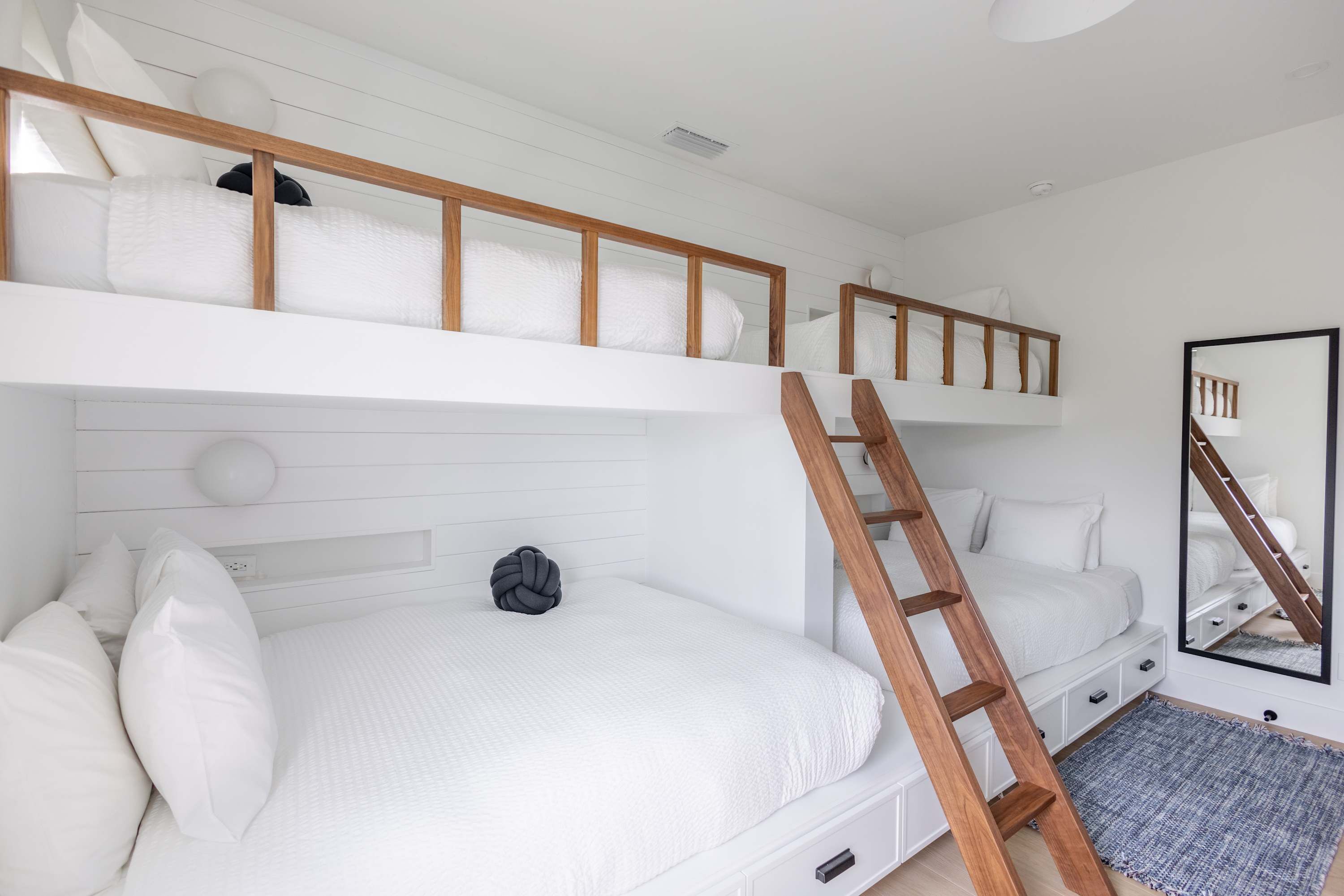 Una cama litera con escalera de madera, ropa de cama blanca y una lámpara redonda pequeña en la pared.