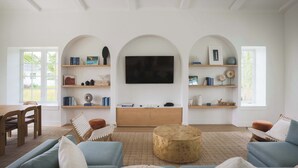 A spacious living room with a large flat-screen TV mounted on the wall, wooden shelves with books and decorative items, a round coffee table, and a seating area with a sofa and armchairs.