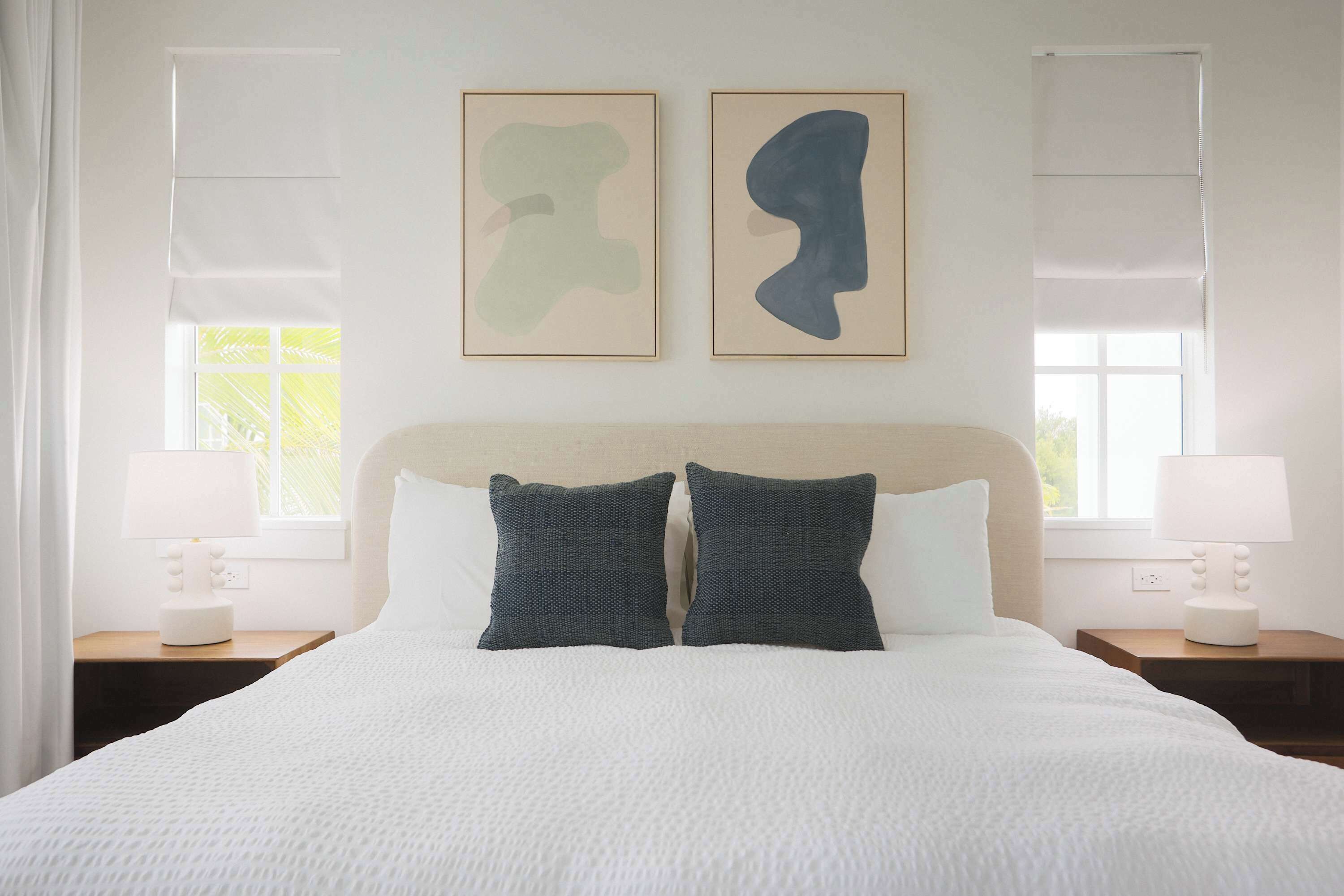 Een net opgemaakt bed met een witte dekbedovertrek, twee donkere kussens en een hoofdbord. Er staan twee nachtkastjes met lampen en er hangt abstracte kunst boven het bed.