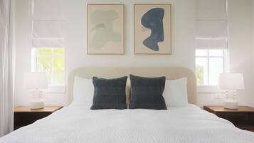 Een net opgemaakt bed met een witte dekbedovertrek, twee donkere kussens en een hoofdbord. Er staan twee nachtkastjes met lampen en er hangt abstracte kunst boven het bed.