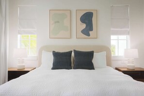 A neatly made bed with a white duvet, two dark pillows, and a headboard. There are two bedside tables with lamps and framed abstract art above the bed.