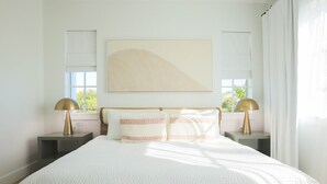 A bedroom with a large bed, two bedside tables with lamps, a window with blinds, and a wall-mounted artwork.
