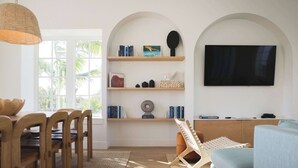 A modern living room with a wooden dining set, a woven light fixture, a flat-screen TV, and a bookshelf with books and decorative items. - Floralee House at Lunara Bay (Key West)