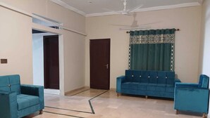 A spacious living room with a blue sofa set, a matching armchair, and a ceiling fan. - Hotel Subhan Palace 2 (Karachi)